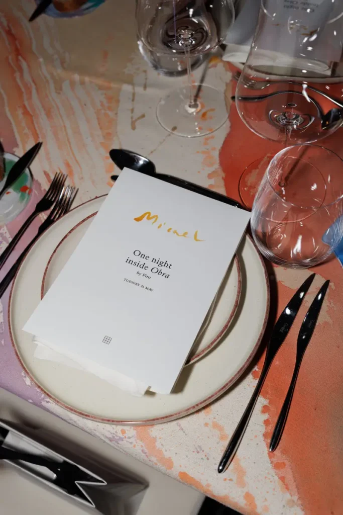 Curated dinner setting with a personalized One Night Inside Obra menu resting atop artist-designed table linens, surrounded by refined glassware and contemporary tableware