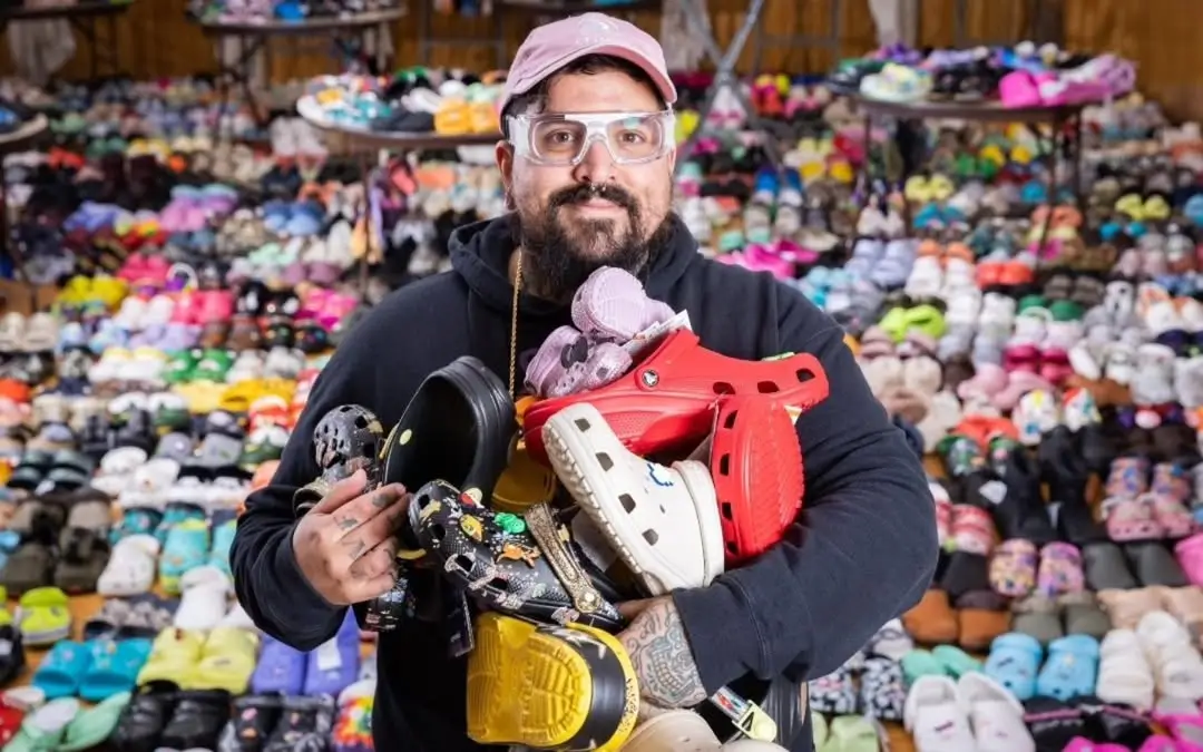 A person stands in front of an expansive Crocs collection while holding an armful of assorted pairs, including bright red, black, cream, and graphic-covered styles. Wearing a cap, clear glasses, and a dark hoodie, they are framed by rows upon rows of colorful Crocs arranged across shelves and the floor, emphasizing the scale and diversity of the collection