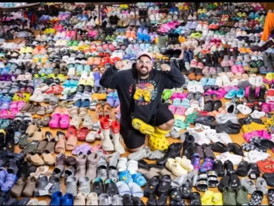 A person kneels and poses in the center of an enormous Crocs collection, surrounded by thousands of pairs arranged across the floor and stacked into the background. The collection features a wide range of colors, silhouettes, collaborations, and themed designs, creating a dense mosaic of footwear that emphasizes scale, variety, and collector culture