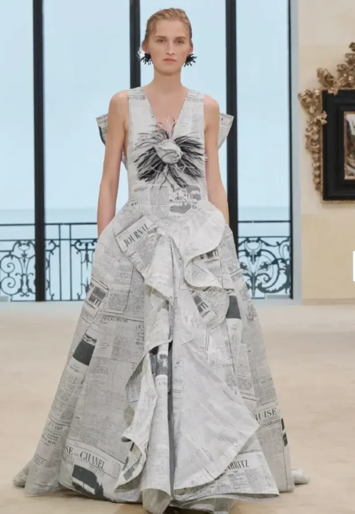 Runway model walking in a structured newspaper-print gown with layered draping and monochrome typography, featuring a sculptural bodice detail and voluminous skirt, set against a bright seaside interior with tall windows and balcony railings