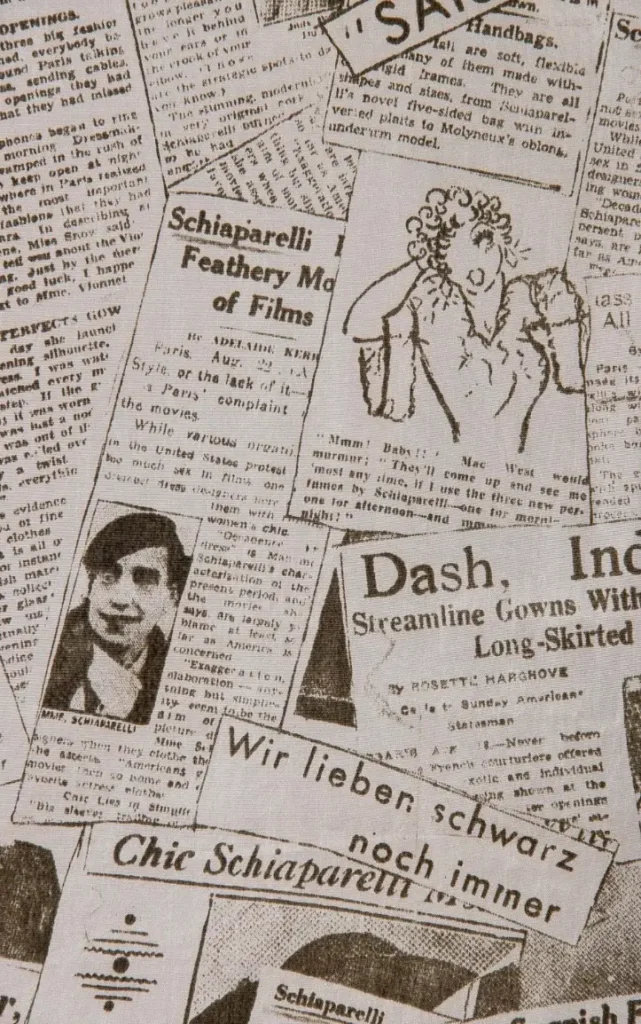 Close-up of monochrome newspaper-print textile featuring layered vintage-style clippings, headlines, and illustrations referencing Schiaparelli, with collage-like typography and archival imagery evoking early 20th-century press culture