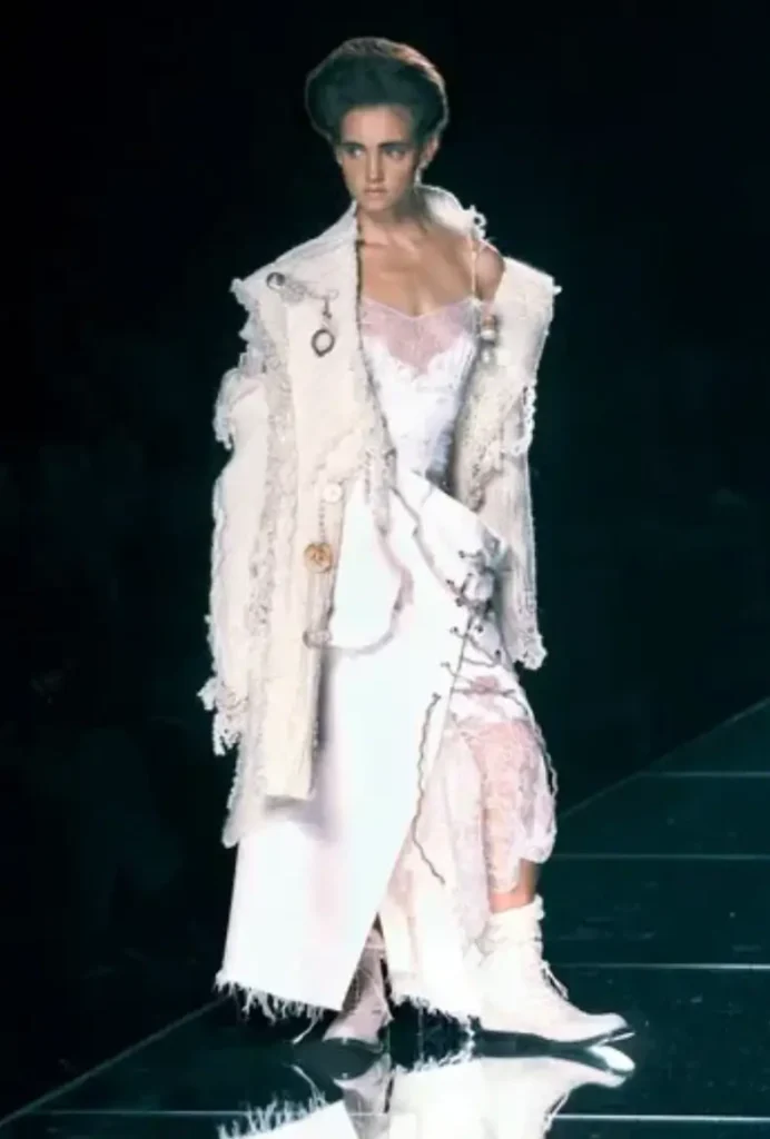 Runway model in layered off-white ensemble featuring textured jacket, lace slip dress, and distressed hems, styled with tall boots and avant-garde hair, walking under dramatic low lighting on a reflective catwalk