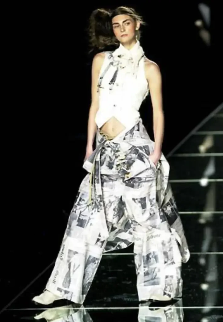 Runway model wearing a tailored white halter vest paired with wide-leg newspaper-print trousers, styled with a neck scarf and hands in pockets, walking under spotlight on a glossy, reflective runway