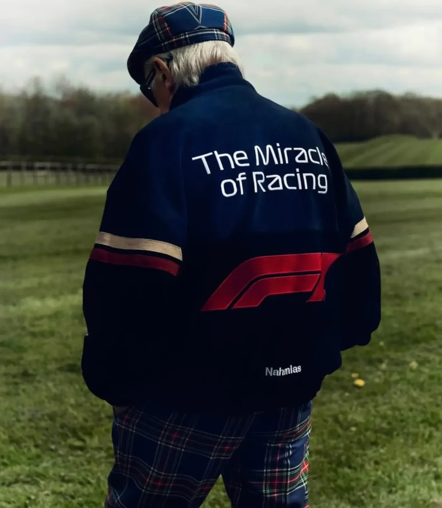 Navy racing jacket worn outdoors, featuring a bold back graphic that centers the phrase “The Miracle of Racing” in clean white lettering. Beneath it, an oversized red Formula 1 logo stretches across the lower back, creating a strong, high-contrast visual against the dark base. Subtle horizontal striping in cream and red runs across the upper arms, reinforcing the motorsport influence. A small “Nahmias” mark sits near the hem, grounding the branding. Styled with a plaid cap and matching trousers, the look blends heritage tailoring cues with modern racing iconography, set against an open, pastoral backdrop that adds a cinematic, lifestyle dimension