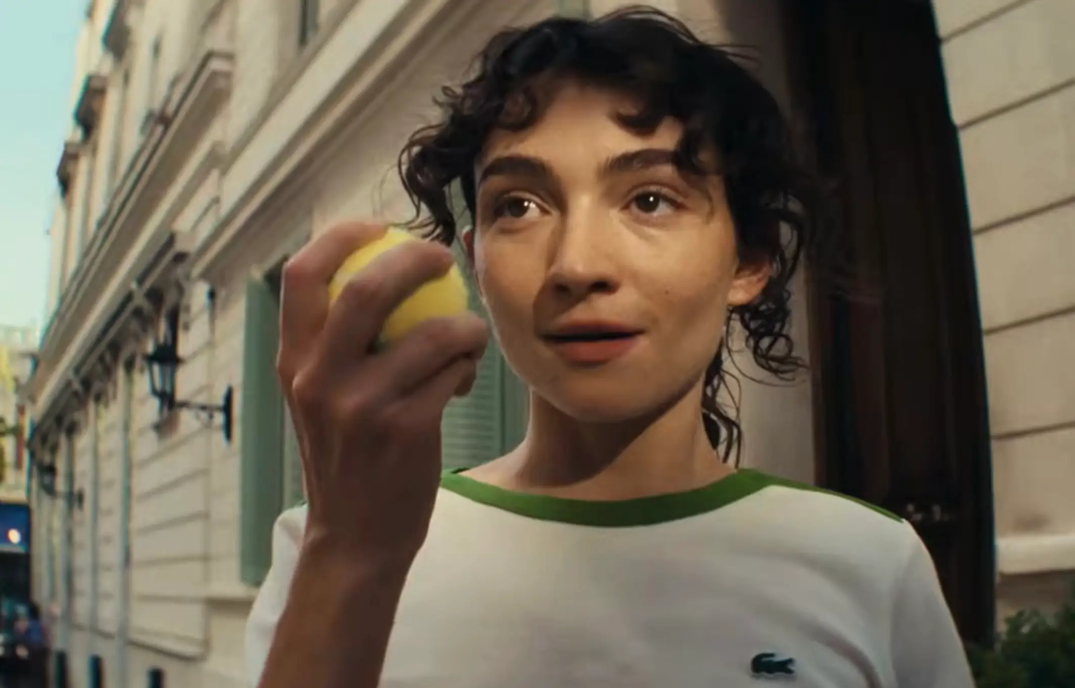 A woman with short, curly hair holds a bright yellow tennis ball up near her face, her expression soft and intrigued. She wears a white top with green trim and stands on a narrow street lined with classic buildings, the warm light and shallow depth of field giving the moment an intimate, cinematic feel