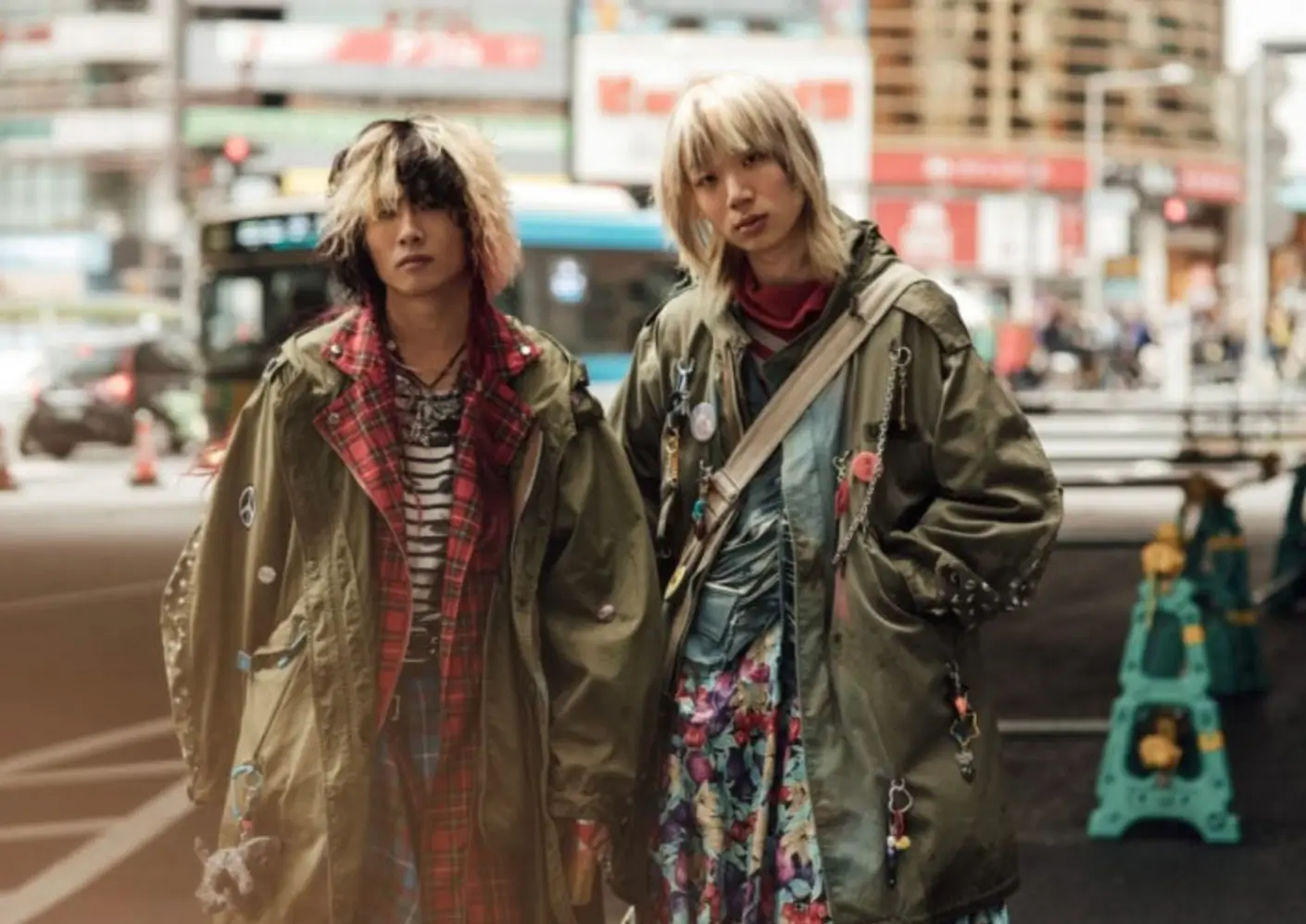 Two figures stand side by side on a busy city street, framed by blurred traffic and signage that suggest an urban Japanese setting. Both wear oversized, military-green coats layered with expressive, almost chaotic styling details. The figure on the left pairs a red tartan layer over a striped top, creating a clash of patterns that feels deliberate rather than accidental. Their hair is heavily textured, split between dark and bleached tones, falling unevenly across the face and reinforcing a raw, DIY aesthetic. Pins and small attachments hang from the coat, adding a sense of personal narrative and customization. The figure on the right contrasts with a softer but equally complex composition. A floral dress flows beneath the coat, introducing movement and color against the otherwise muted outerwear. A crossbody strap cuts diagonally across the torso, while chains, badges, and small objects hang from the jacket, echoing the same language of adornment. Their lighter, layered haircut frames the face with a more refined but still subversive edge. Together, the styling reads as a collision of subcultures—punk, grunge, and streetwear—filtered through a distinctly contemporary lens. The oversized silhouettes, mixed textiles, and accumulation of accessories create a sense of lived-in individuality, where each element feels collected rather than styled. The urban backdrop amplifies this impression, positioning the look within a real, moving environment rather than a staged setting