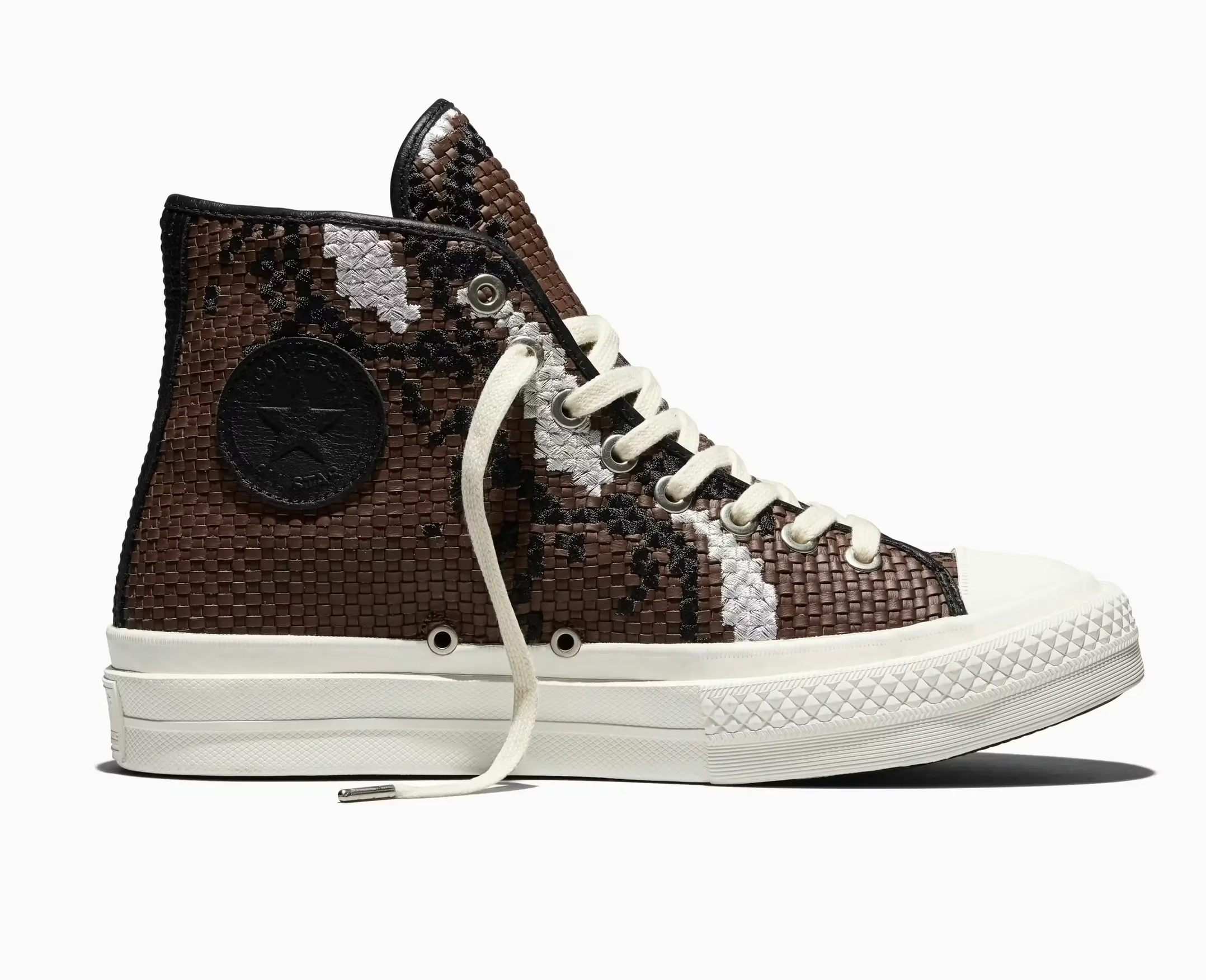 Side profile product shot of the Converse First String Chuck Taylor high-top shoe in brown, featuring an intricately woven leather-style upper with black and metallic silver abstract detailing, tonal black Converse ankle patch branding, cream rope laces, metal eyelets, and a clean off-white vulcanized rubber sole against a light grey studio background