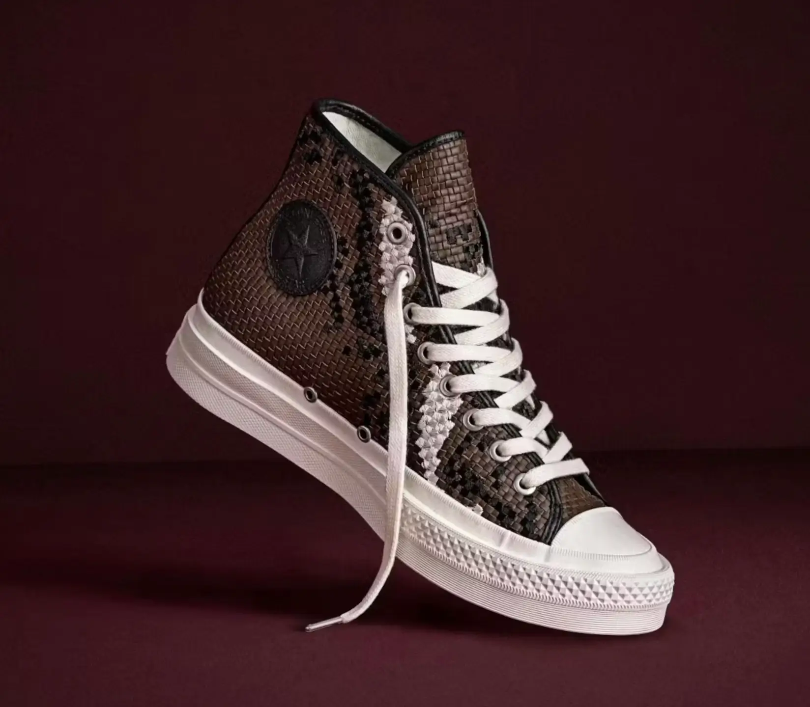 Converse First String Chuck Taylor high-top shoe photographed in a floating studio composition against a deep burgundy backdrop, featuring a woven brown leather-style upper with black and metallic silver abstract detailing, tonal black Converse ankle patch branding, cream rope laces, metal eyelets, and a crisp off-white rubber sole that contrasts the darker textured finish