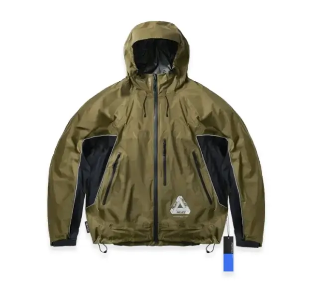Studio product image of a Palace Summer 2026 technical shell jacket in olive green with contrasting black panel inserts, featuring a lightweight hooded construction, multiple waterproof zip pockets, adjustable hem cords, and reflective Tri-Ferg branding near the lower front, presented against a clean light-grey background with retail tag attached