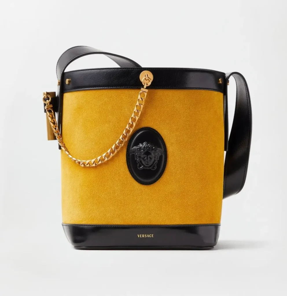 A structured Versace bucket-style shoulder bag is displayed against a light gray studio background, featuring a rich mustard-yellow suede body contrasted with glossy black leather trim and strap details. A chunky gold chain drapes across the front beside a black Medusa plaque emblem, while subtle gold hardware and minimalist branding at the base reinforce the bag’s blend of archival glamour and contemporary luxury craftsmanship