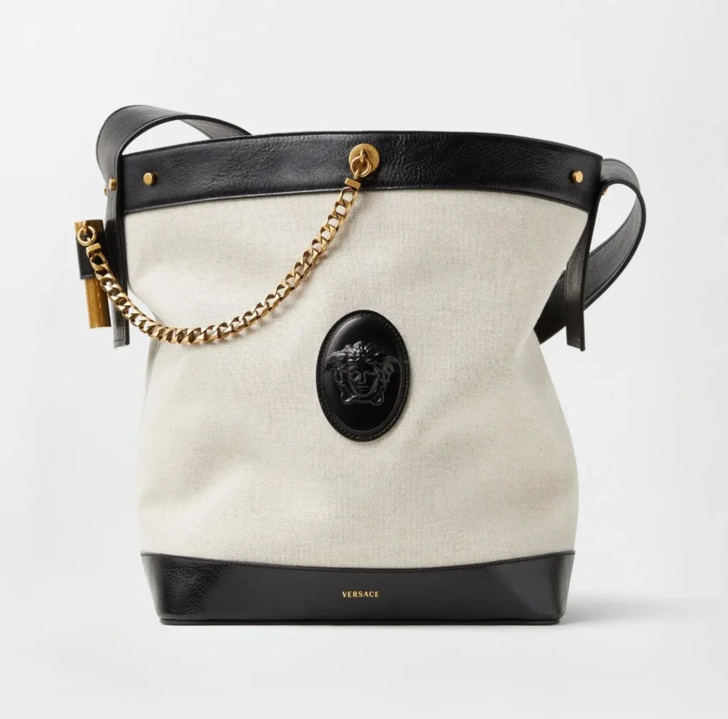 A Versace bucket-style shoulder bag is presented against a minimalist light gray studio backdrop, combining an off-white canvas body with glossy black leather trim and strap detailing. The design features a black Medusa plaque emblem centered on the front, accented by a chunky gold chain and polished gold hardware, while the softly structured silhouette blends relaxed resort sensibility with refined Italian luxury craftsmanship