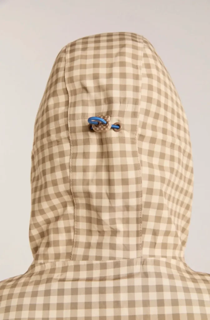 A close-up rear view of a hooded gingham jacket in soft beige and cream tones, highlighting the garment’s technical construction and subtle detailing. The hood features paneled stitching with a central adjustable toggle system threaded through vivid cobalt-blue cord loops, creating a sharp visual contrast against the muted check pattern. Gentle fabric creasing and precise seam lines emphasize the lightweight outerwear fabrication, while the enlarged crop draws attention to the balance between utilitarian function and refined contemporary styling
