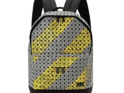 Front-facing view of the Bao Bao Issey Miyake Daypack Autobahn backpack in gray and yellow, featuring the brand’s signature triangular geometric panel construction. Bold diagonal yellow stripe graphics cut across the reflective gray surface, while black padded shoulder straps and a reinforced black base emphasize its industrial, urban-inspired design language