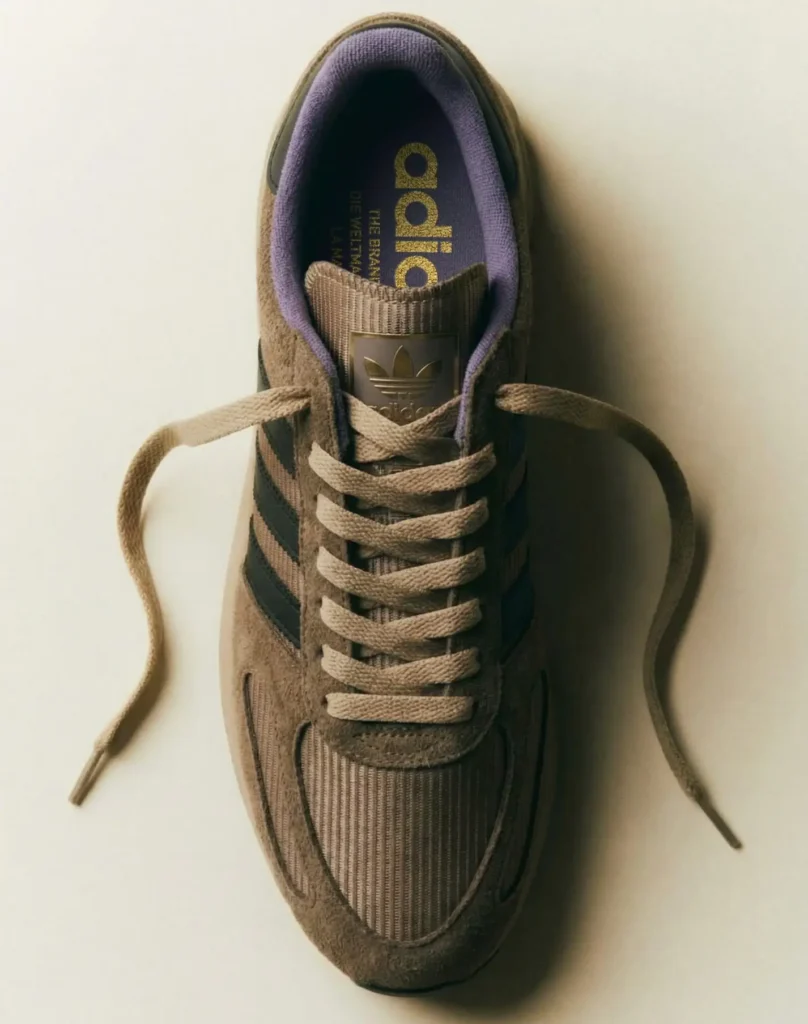 A top-down studio image highlights the adidas L.A. Trainer in earthy brown suede and textile materials with tonal laces, black Three Stripes branding, and a soft purple inner lining, emphasizing the sneaker’s retro athletic identity through a refined contemporary presentation