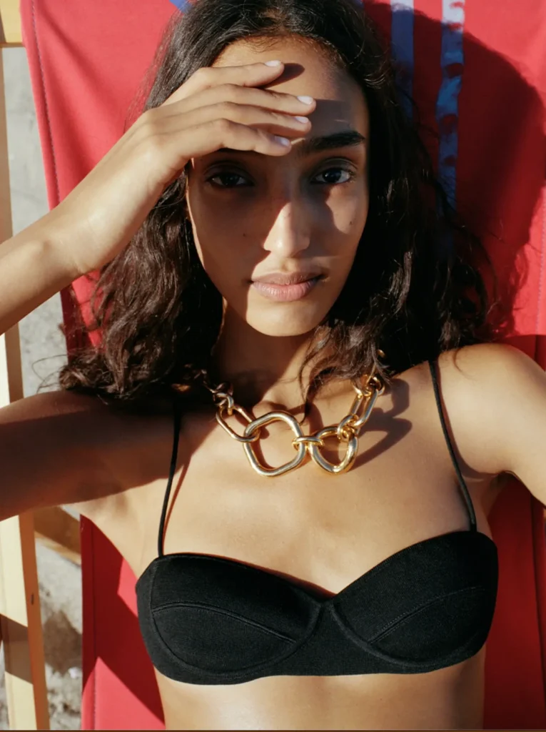 A model reclines on a red lounge chair under strong afternoon sunlight, wearing a minimalist black bikini top with thin straps and a chunky sculptural gold chain necklace. Her hand partially shields her face from the sun, creating dramatic shadows across her features, while the warm lighting and close-cropped composition give the image a refined luxury-fashion editorial fee