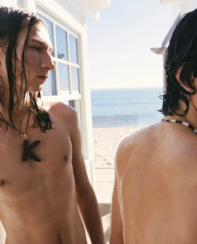 Two wet-haired models stand near a beachside cabana in bright coastal sunlight, their skin still damp from the water. One faces sideways wearing a bold oversized “K” pendant necklace, while the other turns away from the camera, creating a cropped, intimate composition against a backdrop of pale sand, calm ocean water, and soft summer haze