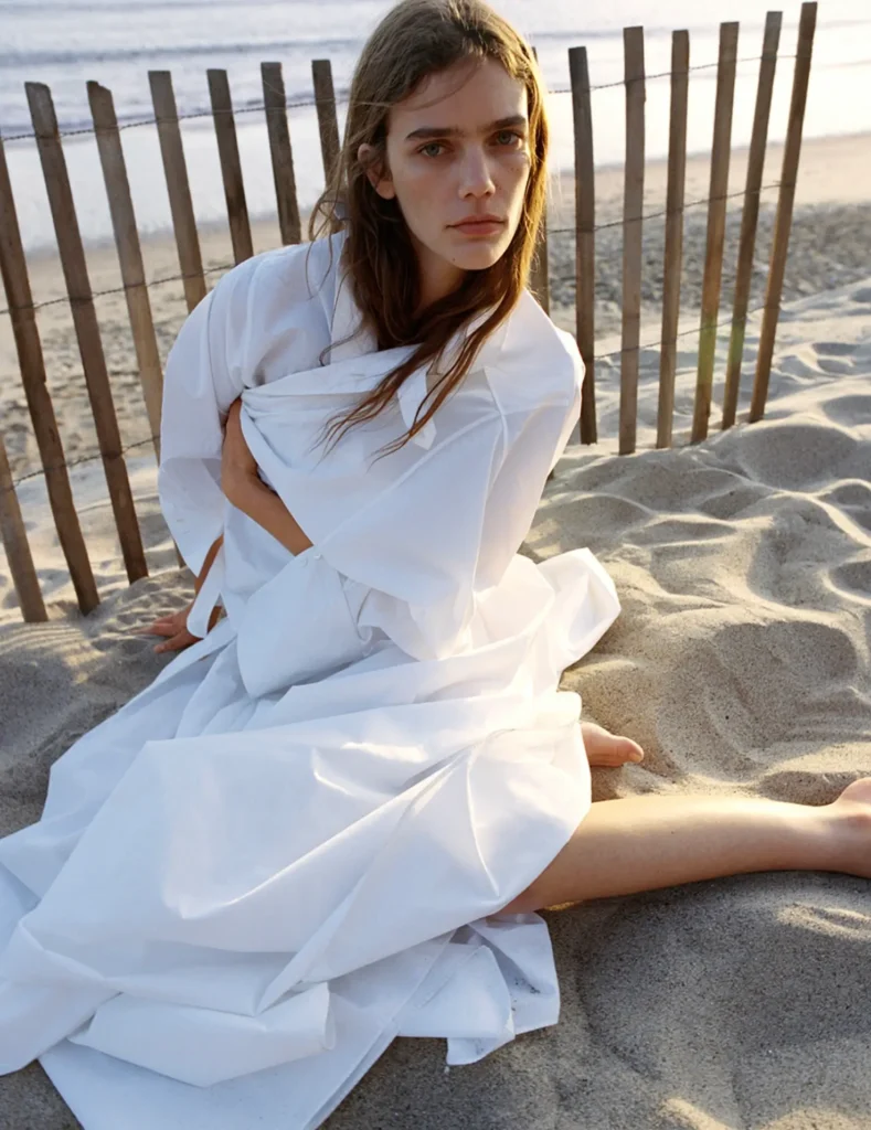 A model sits barefoot in the sand beside a weathered beach fence, wrapped in an oversized flowing white shirt dress illuminated by soft golden-hour light. The relaxed pose, windswept hair, and muted coastal backdrop create a calm, cinematic fashion editorial atmosphere rooted in minimalism and quiet