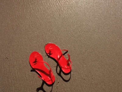 A pair of vivid red thong sandals rests on wet shoreline sand beneath direct sunlight, casting sharp elongated shadows across the textured surface. The minimalist composition and saturated color contrast create a clean, cinematic luxury-fashion still life with a summery coastal mood