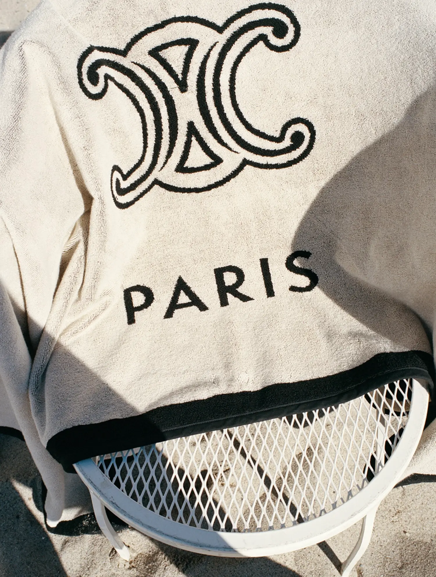 A cream-colored beach towel featuring a bold black “Paris” logo and ornate monogram graphic is draped over a white mesh chair on the sand. Warm sunlight and deep shadows emphasize the textured fabric and minimalist coastal setting, giving the image a refined haute-resort editorial feel