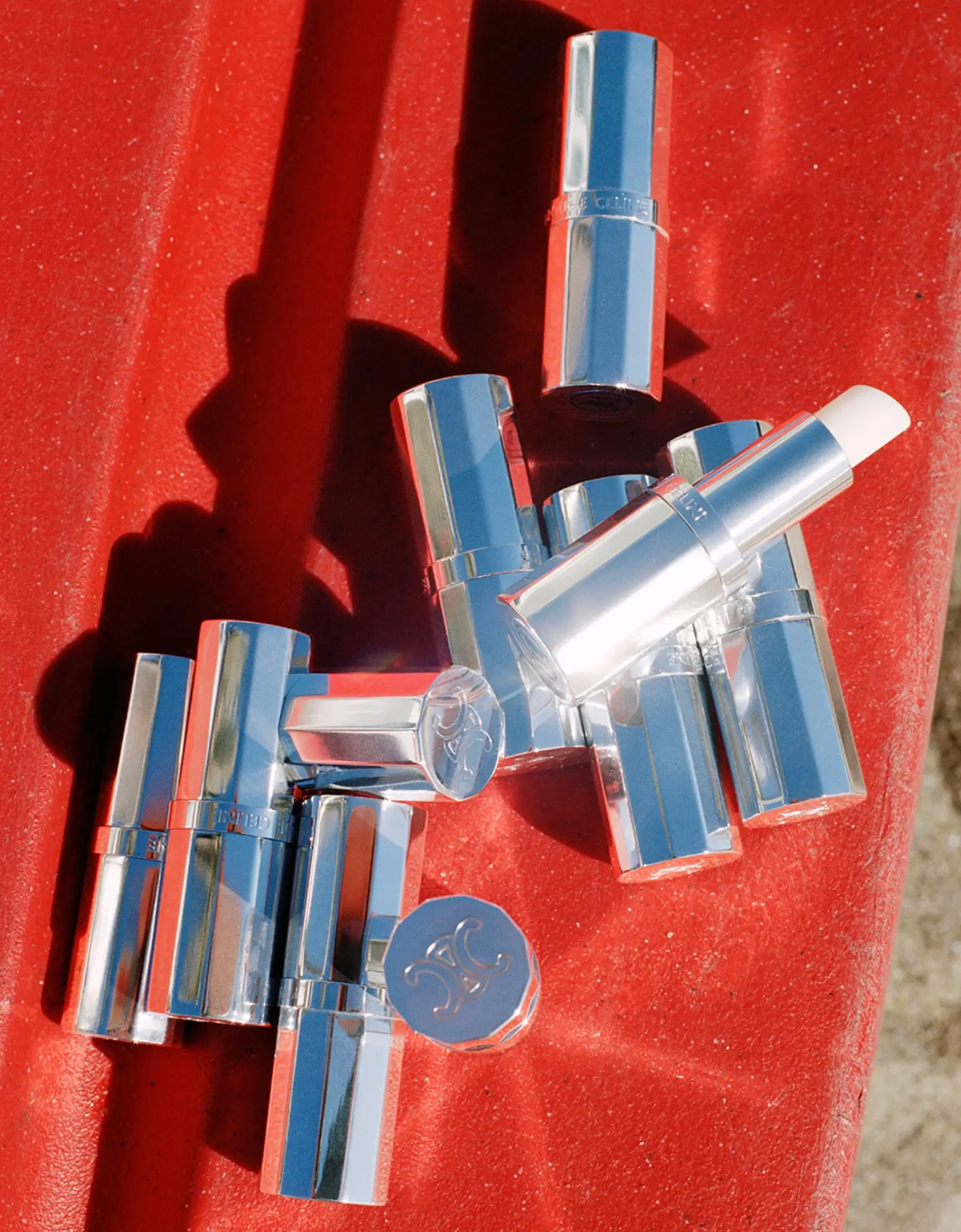 Several polished silver lipstick and balm cases are scattered across a vivid red lounge surface under intense sunlight, casting dramatic elongated shadows. The reflective metallic packaging and saturated color contrast create a sharp, high-gloss beauty editorial with a luxurious summer-resort aesthetic