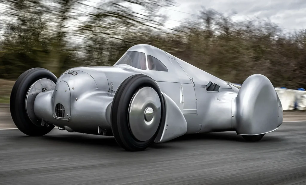 A streamlined silver coachbuilt race car speeds along a roadway, its sculptural aluminum bodywork shaped with dramatic teardrop curves, enclosed rear wheels, and exposed front wheel arches. The vehicle’s riveted metal panels, narrow cockpit canopy, and pre-war aerodynamic proportions evoke vintage land-speed record machines, while the motion-blurred forest backdrop reinforces its sense of velocity and handcrafted mechanical elegance