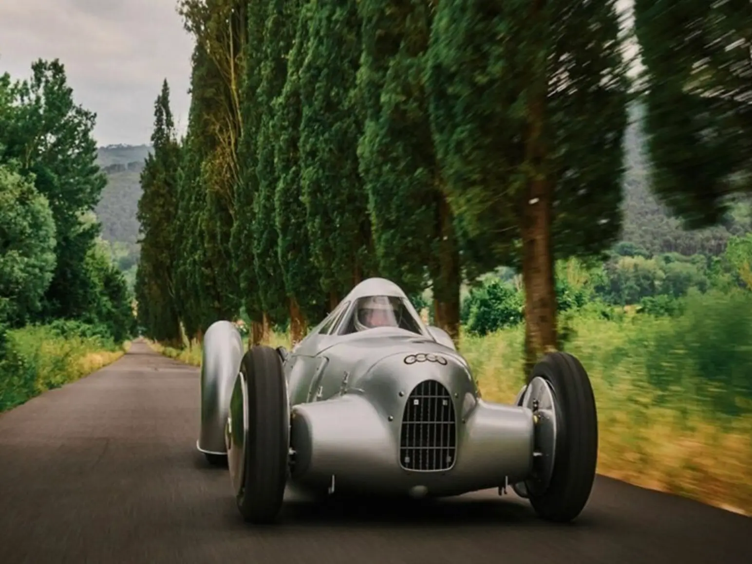 A polished silver Auto Union streamliner speeds down a narrow avenue lined with towering cypress trees, combining pre-war Grand Prix proportions with aircraft-inspired aerodynamic bodywork. The car’s exposed front wheels, enclosed rear fenders, riveted aluminum panels, and compact canopy cockpit create a dramatic Silver Arrow silhouette, while the motion-blurred countryside emphasizes the machine’s historic obsession with speed and engineering innovation