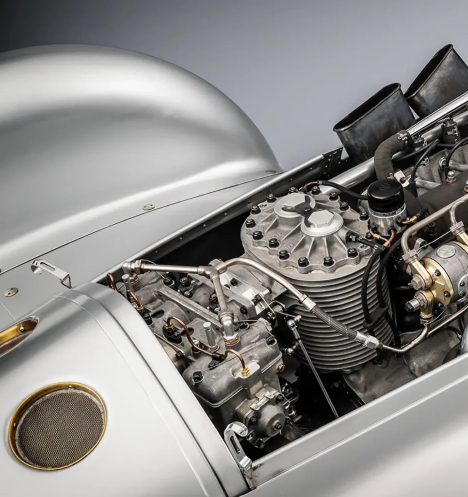 A close-up detail view reveals the exposed mechanical heart of a handcrafted silver streamliner race car, showcasing a meticulously engineered vintage engine surrounded by polished aluminum bodywork. Ribbed metal casings, visible fuel and oil lines, intake trumpets, riveted panels, and brass-trimmed ventilation elements emphasize the machine’s fusion of pre-war motorsport engineering, aviation-inspired craftsmanship, and sculptural industrial design