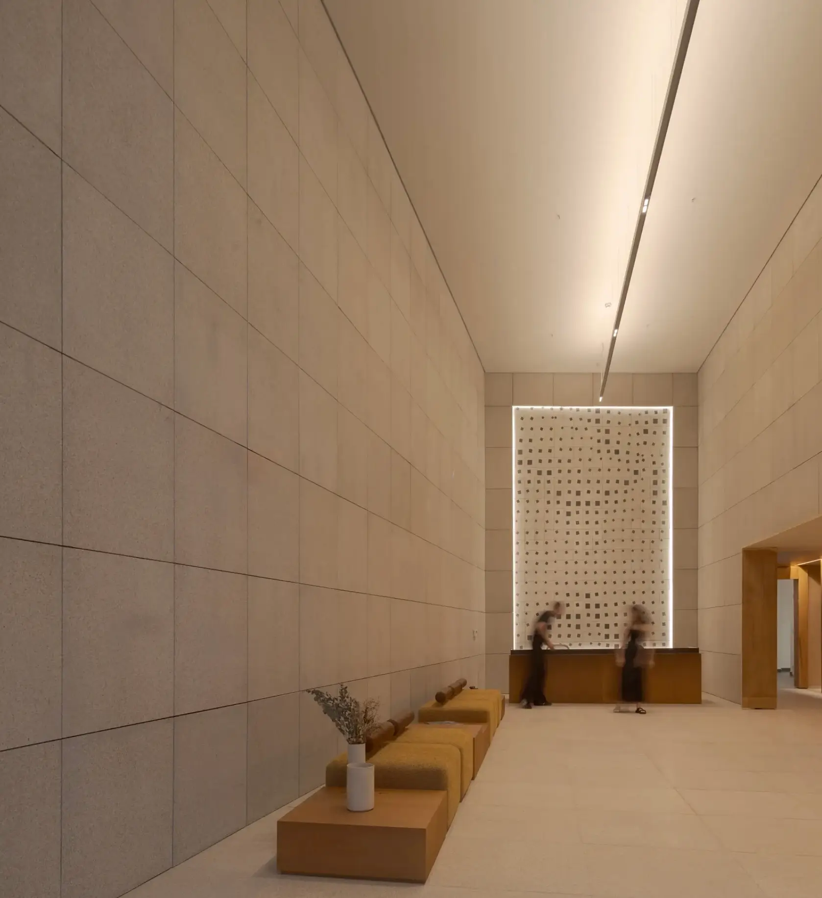 A monumental minimalist lobby interior features soaring stone-clad walls, recessed linear ceiling lighting, and a restrained palette of warm beige and sand tones. Low modular seating lines one side of the elongated space, leading toward an illuminated perforated wall installation behind a reception desk, while blurred figures introduce movement and scale within the serene contemporary architectural environment