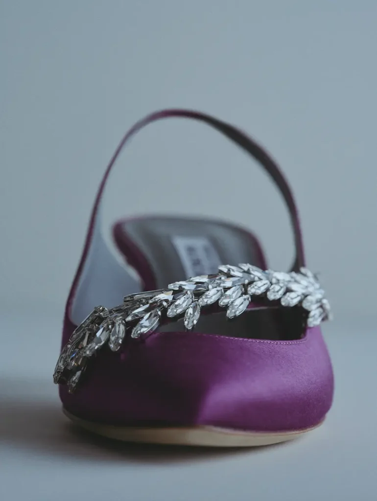 Close-up editorial photograph of a jewel-embellished satin mule in rich violet-purple. The pointed-toe silhouette is finished with an oversized crystal leaf motif across the front, catching soft directional light. A second shoe appears blurred in the background, creating depth and emphasizing the couture craftsmanship and polished eveningwear aesthetic
