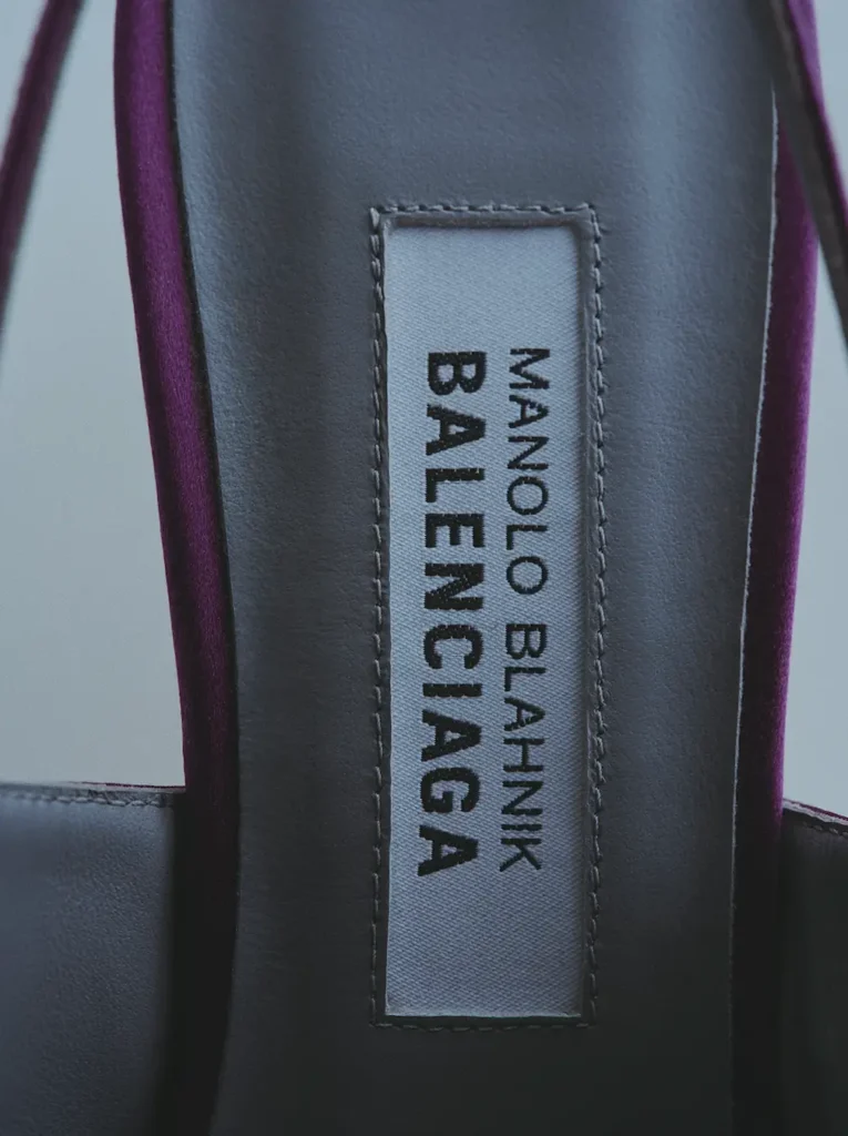 Extreme close-up of the interior branding on a luxury satin mule from the Manolo Blahnik x Balenciaga collaboration. The image highlights the stitched grey insole label with dual-brand typography framed by smooth black leather lining and deep plum satin edges, emphasizing craftsmanship, material contrast, and couture-level finishing details