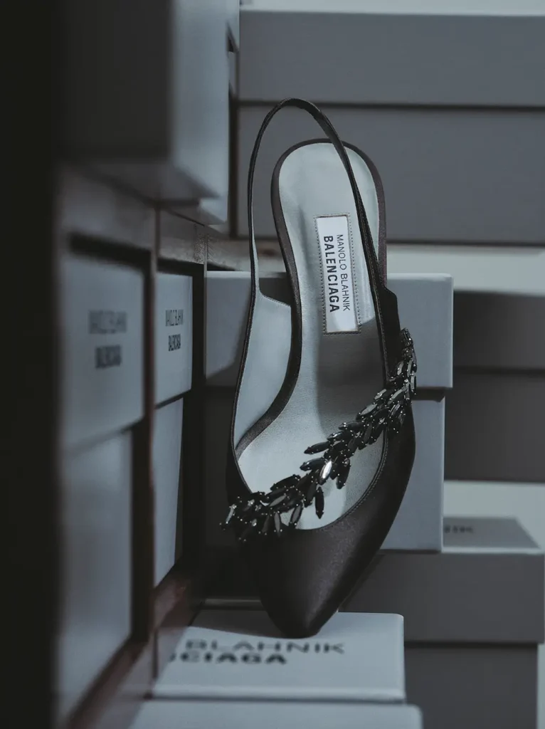 Editorial product photograph of a black satin slingback heel from the Manolo Blahnik x Balenciaga collaboration, positioned among stacked branded shoe boxes. The pointed silhouette is accented with an oversized black crystal leaf embellishment across the vamp, while the co-branded insole label remains visible inside. Soft sils, muted tones, and narrow framing create a dramatic luxury archive and atelier display aesthetic