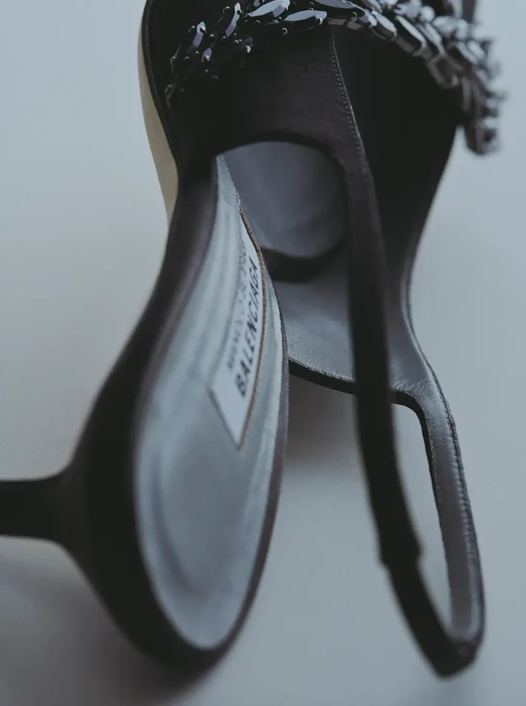 rtistic close-up detail of a black satin slingback heel from the Manolo Blahnik x Balenciaga collaboration, photographed from an angled interior perspective. The image emphasizes the sculpted opening, smooth grey leather insole, slim slingback strap, and partially visible co-branded label, while glossy black crystal embellishments frame the upper edge. Soft focus and muted lighting create an intimate couture craftsmanship aesthetic