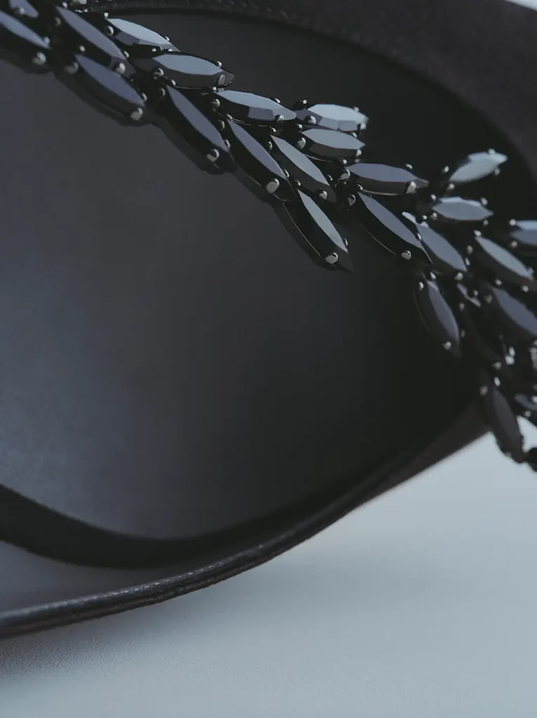 Extreme close-up editorial detail of a black satin luxury shoe featuring an intricate black crystal embellishment across the upper. The elongated marquise-cut stones are arranged in a leaf-like formation, creating tonal texture and subtle shine against the smooth satin surface. Soft directional lighting and shallow depth of field emphasize couture craftsmanship and material contrast