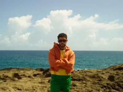 A man stands on a rugged coastal landscape with the ocean and towering clouds behind him, wearing an orange zip hoodie layered over a bright yellow shirt and green bottoms. Dark sunglasses and folded arms reinforce the relaxed yet self-assured mood, while the saturated colors create a playful, sun-drenched island aesthetic