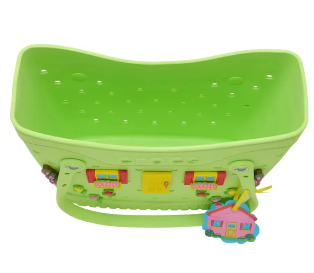 Lime green Crocs tote bag designed like a miniature house façade, featuring molded windows with flower boxes, a bright yellow front door, floral details, and a dangling pink house-shaped charm. The bag has a structured silhouette with perforated detailing inside and playful pastel cottage-inspired accents throughout