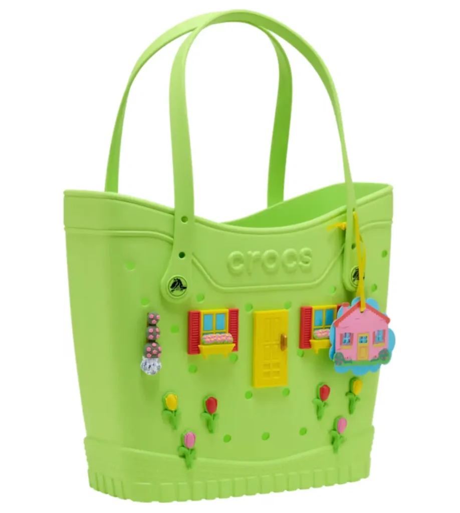 Front view of a lime green Crocs tote bag styled as a whimsical cottage, featuring miniature windows with red shutters, flower boxes, a bright yellow front door, scattered tulip embellishments, and a dangling pink house-shaped charm. The structured bag includes long shoulder straps and subtle perforated detailing inspired by classic Crocs design lang