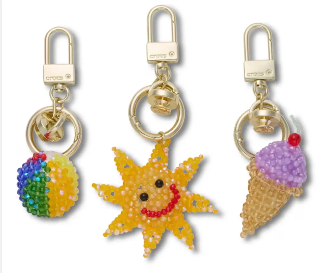 Set of play beaded keychain charms attached to gold-tone Crocs clips, featuring a rainbow beach ball, a smiling yellow sun with a red smile, and a lavender ice cream scoop in a waffle cone. Each charm is handcrafted with faceted beads and designed with a whimsical, summery aesthetic