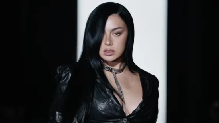 Close-up still from the “SS26” music video featuring Charli XCX in a sleek black look with a deep neckline, long dark hair partially covering one eye, and a metallic chain necklace, framed against dramatic monochrome runway-style lighting