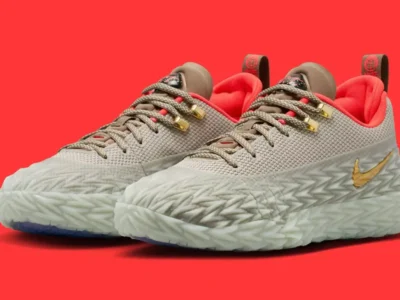 Angled front three-quarter view of the Nike ST Charge “College Grey” basketball shoes featuring a grey engineered mesh upper, sculpted translucent armored wrap, metallic gold Swoosh and lace hardware, bright crimson inner lining, and icy blue outsole accents designed for outdoor court performance