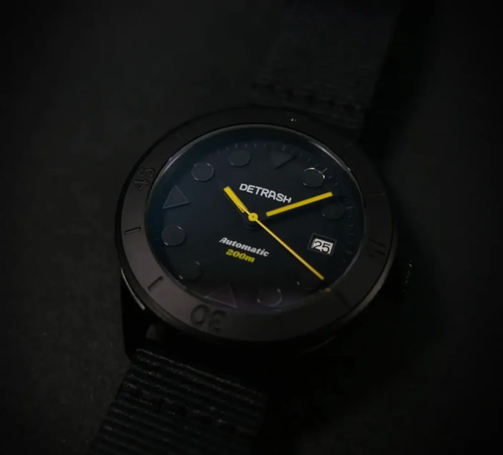 Moody angled product photograph of the Detrash Gotham’s Vigilante 2 automatic dive watch resting in near darkness. The stealth-black dial and bezel blend into the sil while vivid yellow hands create sharp contrast, emphasizing the Batman-inspired visual identity. Circular and triangular markers, the framed date window, and textured recycled strap reinforce the watch’s minimalist tactical aesthetic and nocturnal character