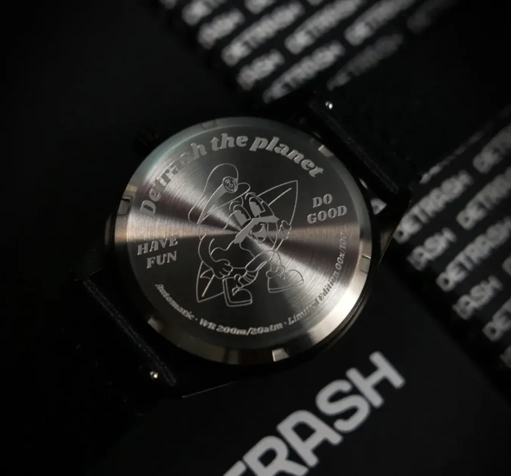 Close-up product photograph of the Detrash Gotham’s Vigilante 2 caseback showcasing an engraved illustration and the brand’s playful sustainability messaging. The brushed metal back features phrases including “Detrash the planet,” “Have Fun,” and “Do Good,” alongside technical markings and limited-edition numbering. Set against a dark, cinematic backdrop, the image highlights the watch’s blend of eco-conscious storytelling, collectible character, and stealth-inspired design language