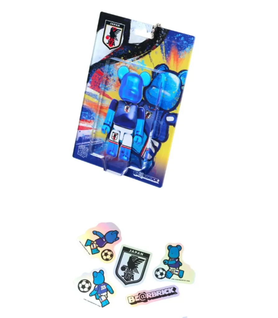 Official BE@RBRICK × Japan National Soccer Team (SAMURAI BLUE) accessory set featuring a packaged translucent blue mini BE@RBRICK figure against dynamic football-inspired artwork alongside holographic stickers showing BE@RBRICK characters, football graphics, the JFA crest, and connective branding elements