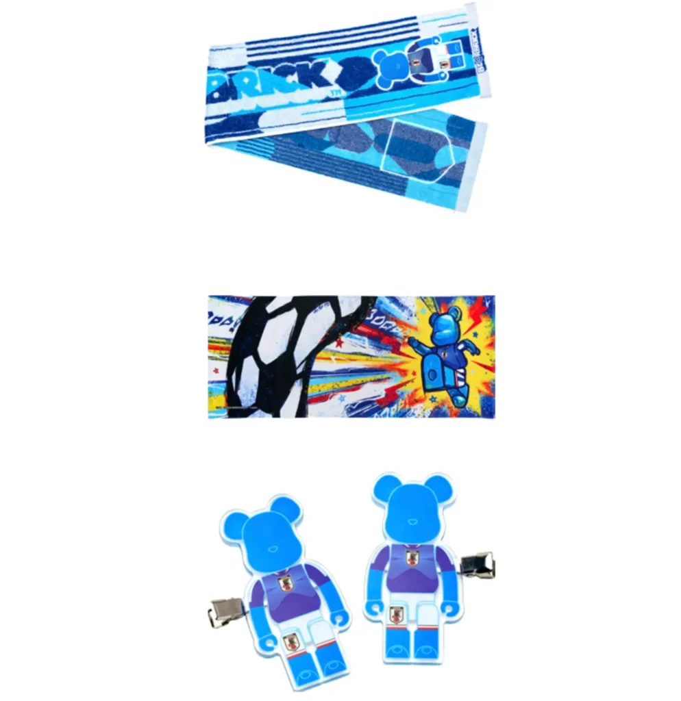 Selection of official BE@RBRICK × Japan National Soccer Team (SAMURAI BLUE) accessories including a blue football supporter towel with BE@RBRICK graphics, an illustrated sports-themed artwork card featuring a bear figure and football motion graphics, and a pair of translucent blue BE@RBRICK character clips styled in Japan’s national team kit with JFA crest detail