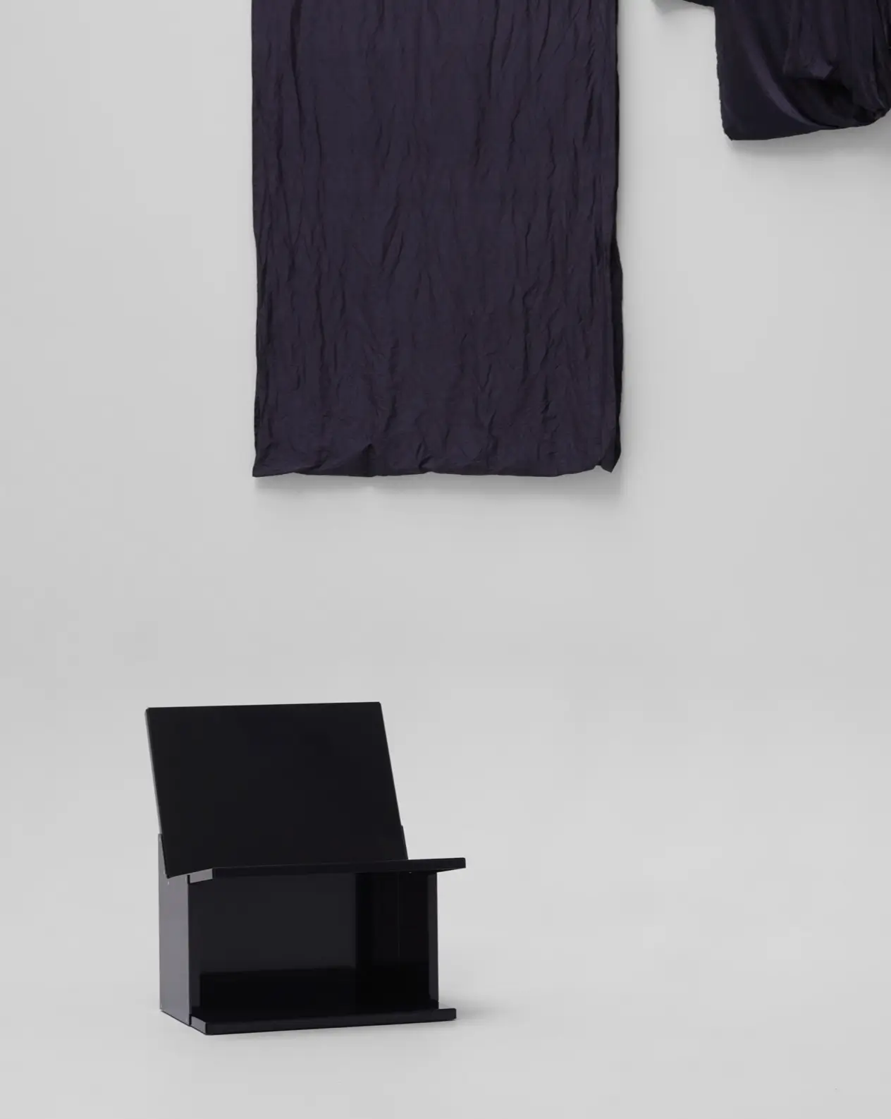 Minimalist black sculptural lounge chair with an integrated open storage base displayed in a gallery-like interior, positioned beneath suspended dark textile panels that emphasize its architectural form, restrained proportions, and contemporary design aesthetic