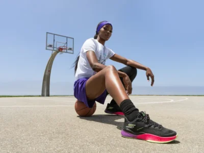 Rickea Jackson poses on an outdoor basketball court wearing black and neon-accented Skechers Basketball performance sneakers, paired with purple athletic shorts and a white graphic tee. Sitting confidently on a basketball beneath a clear blue sky, the scene emphasizes the growing connection between women’s basketball culture, performance footwear innovation, and modern athlete-driven style