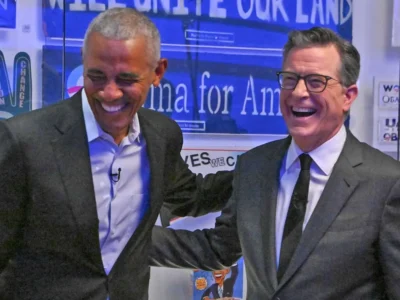 Barack Obama and Stephen Colbert share a candid laugh backstage during a campaign-era appearance, surrounded by Obama-themed posters, signs, and memorabilia. The image captures the long-running rapport between the former president and late-night host, blending political history with the relaxed energy that has defined many of their televised conversations