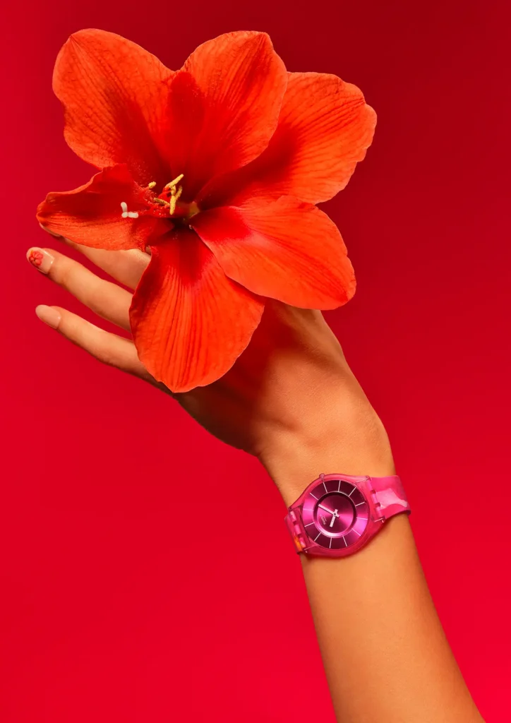 A vibrant editorial-style close-up captures one of Swatch’s PAINTED PARADISE SKIN models against a saturated crimson backdrop, emphasizing the collection’s tropical escapist energy through bold monochromatic color play. The ultra-thin translucent pink watch wraps around the wrist with feather-light minimalism, while its rich magenta dial radiates subtle metallic depth through concentric detailing. A large coral-red tropical flower rests delicately in the model’s hand, visually echoing the watercolor-inspired tones and floral influences behind the collection’s paradise-driven aesthetic. The composition balances softness and intensity simultaneously, merging fashion photography, botanical symbolism, and Swatch’s playful Swiss design language into a striking summer-ready campaign view