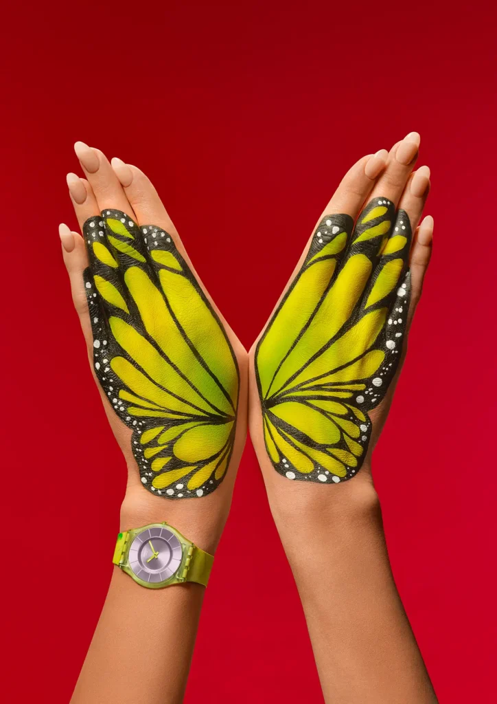 A vivid editorial campaign image merges tropical color theory and body art through a striking butterfly-wing composition painted across a model’s hands against a saturated crimson-red backdrop. The symmetrical yellow-and-black wing illustration evokes monarch butterfly patterns, with bold graphic outlines and white dotted detailing creating sharp visual contrast against the warm skin tones and hyper-saturated background. Anchoring the composition is a translucent lime-green Swatch PAINTED PARADISE SKIN watch featuring a soft metallic lavender dial and watercolor-inspired marbled strap textures that subtly echo the organic movement of the painted wings. The ultra-thin minimalist silhouette enhances the image’s contemporary fashion-editorial aesthetic, while crisp studio lighting sharpens every contour, texture, and painted detail. The composition transforms the watch into part of a larger surreal tropical fantasy, reinforcing Swatch’s playful fusion of Swiss minimalism, wearable art, and escapist summer expression through color, nature symbolism, and bold graphic storytelling