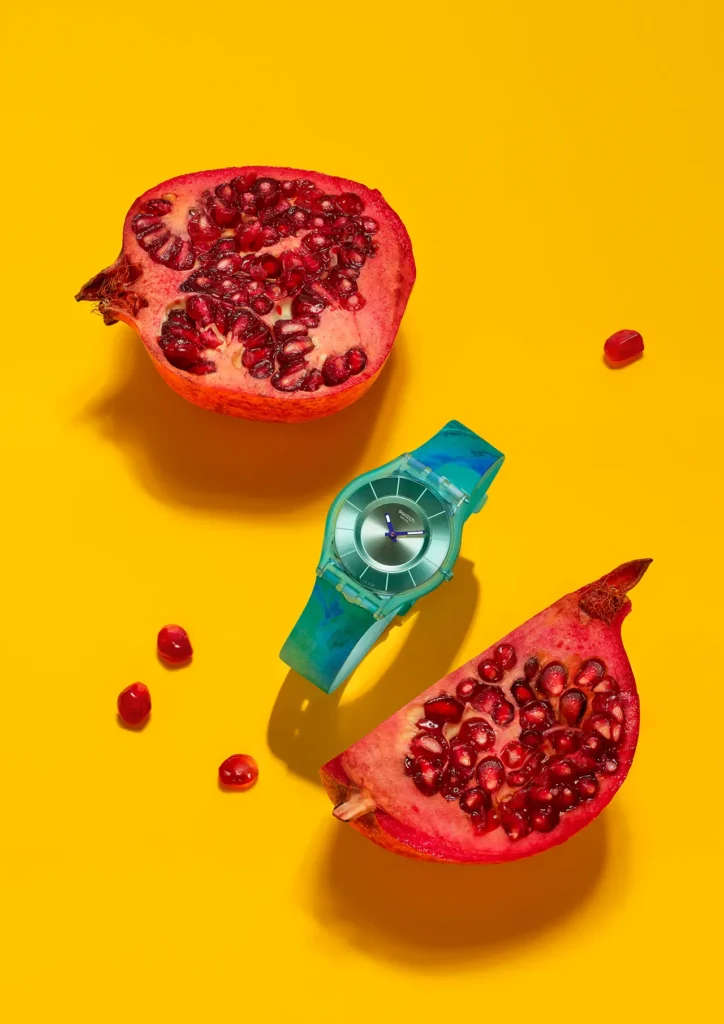 A vibrant still-life campaign image spotlights one of Swatch’s PAINTED PARADISE SKIN watches surrounded by freshly sliced pomegranates against an electrifying sunflower-yellow backdrop. The ultra-thin translucent turquoise watch sits diagonally across the composition, its cool aquatic tones contrasting sharply against the saturated reds of the exposed fruit and scattered ruby seeds. Watercolor-inspired marbling across the strap reinforces the collection’s tropical, painterly aesthetic, while the metallic dial introduces a sleek minimalist balance to the otherwise explosive color palette. Strong directional lighting creates dramatic shadows beneath the fruit and watch, giving the image a graphic editorial sharpness that feels simultaneously playful, contemporary, and fashion-forward. The composition channels Swatch’s signature blend of Swiss minimalism and joyful escapism, transforming everyday objects into a bold visual narrative centered on tincture, lightness, and sensory summer energy