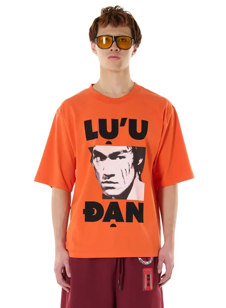 A model wears an oversized neon-orange graphic tee from the LỰU ĐẠN x Bruce Lee collaboration featuring a stark black-and-white portrait of Bruce Lee framed by bold “LU’U ĐẠN” typography. Styled with burgundy athletic shorts accented by martial arts-inspired patches and amber-tinted oversized sunglasses, the look channels the capsule’s fusion of kung fu cinema iconography, rebellious streetwear energy, and retro graphic design. The vivid orange palette and oversized silhouette amplify the collection’s explosive “dangerous man” aesthetic with unapologetic view intensity